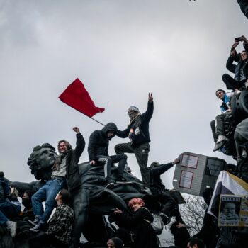 People stand atop a statue in protest holding a red flag.